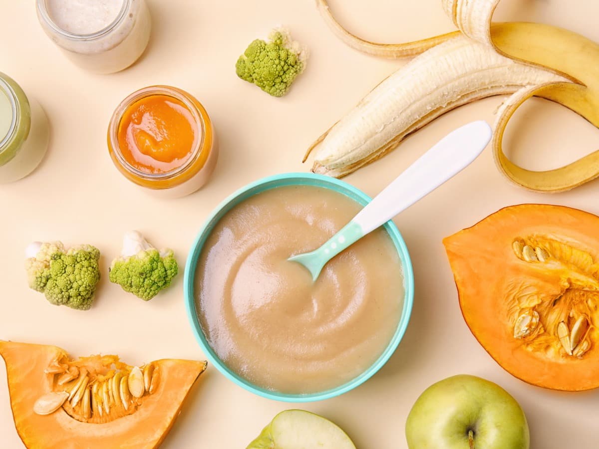 Eine Schale Babybrei, daneben Obst, Gemüse und kleine Gläschen